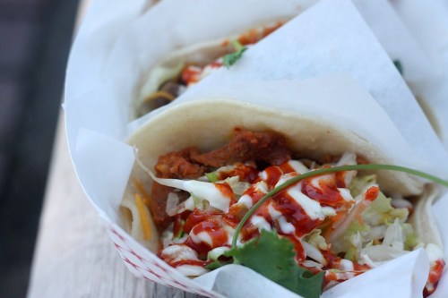 Close-up of tacos in a basket, filled with meat, lettuce, and drizzled with sauce, garnished with herbs.