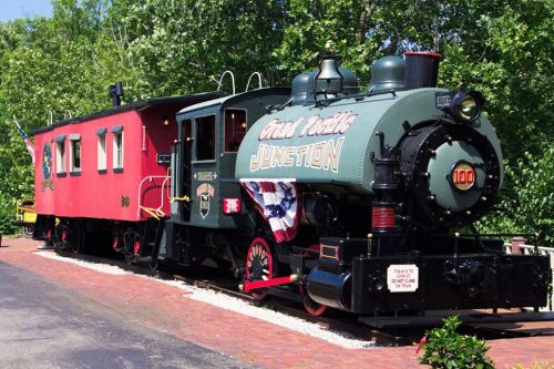A vintage steam locomotive with a red caboose, displayed outdoors among green trees.
