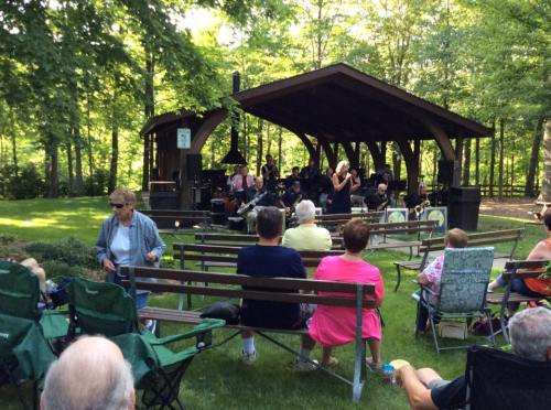 A live band performs under a wooden pavilion as an audience enjoys the music in a lush, green park setting.