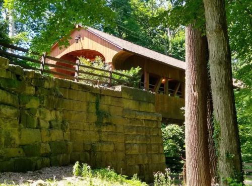A wooden covered bridge nestled among trees, supported by a stone wall, in a lush green setting.