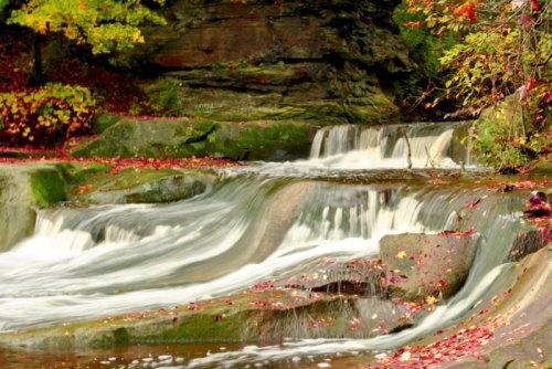 A serene waterfall cascades over rocks, surrounded by autumn leaves in vibrant colors.