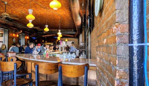 A cozy bar with a brick wall, wooden chairs, and patrons enjoying drinks under warm pendant lights.