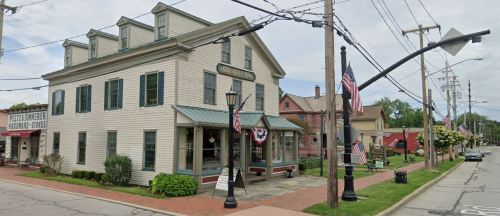 Historic building with green roof and large windows, surrounded by trees and flags, on a quiet street.