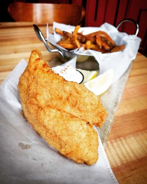 A golden-brown fried fish fillet served with fries, lemon wedges, and a side of dipping sauce on a wooden table.