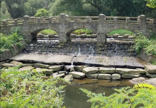 A stone bridge arches over a small waterfall, surrounded by lush greenery and rocky riverbanks.