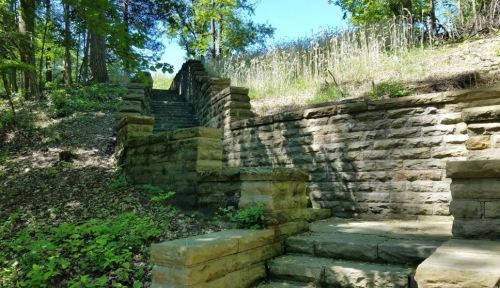 Stone steps lead up a hillside, surrounded by greenery and trees under a clear blue sky.