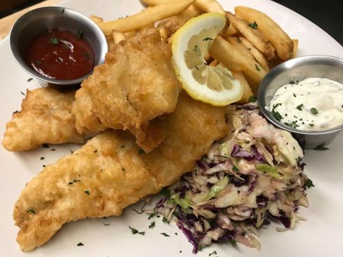 A plate of crispy fried fish fillets, coleslaw, French fries, and dipping sauces, garnished with a lemon slice.