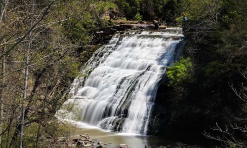 A cascading waterfall surrounded by lush greenery and rocky terrain, with a person standing nearby.