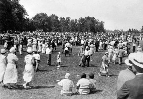 A large crowd of people in vintage clothing gathered in a park on a sunny day, with trees in the background.