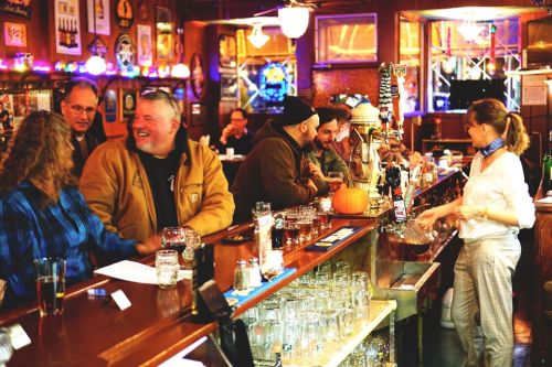 A lively bar scene with patrons chatting at the counter, a bartender serving drinks, and a pumpkin decoration on the bar.