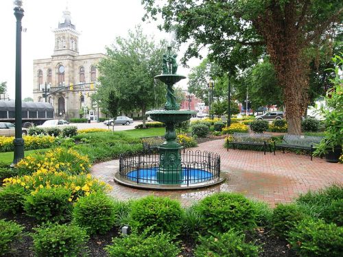 A decorative fountain surrounded by flowers and greenery in a park, with a historic building in the background.