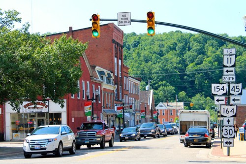 A street scene with traffic lights, shops, and cars in a small town, surrounded by green hills.