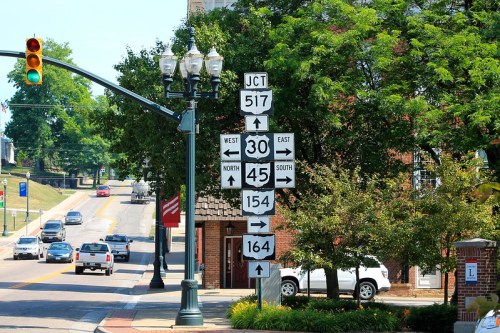 Traffic signs at an intersection showing directions for multiple routes, with vehicles and greenery in the background.
