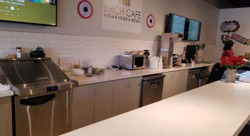 A clean café counter with appliances, plants, and a staff member preparing food in a vegan café setting.