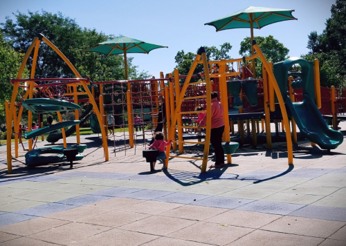 A playground with colorful equipment; a child plays while an adult supervises nearby. Sunny day with green trees.