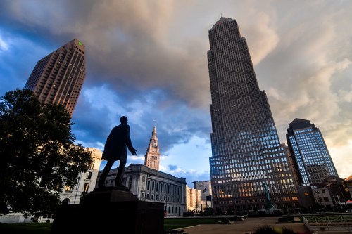 Silhouette of a statue in front of tall skyscrapers under a dramatic sky with clouds.