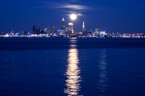 Cleveland skyline at night with a full moon reflecting on the water. City lights twinkle against a dark sky.