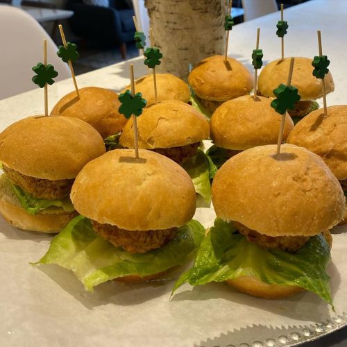 A platter of small sandwiches with lettuce and chicken patties, topped with green clover toothpicks.