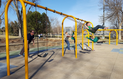 A playground scene with children swinging and playing, while an adult pushes one child on a swing.