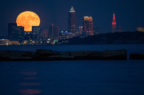 A large full moon rises over the Cleveland skyline, illuminating the city against a dark blue sky.