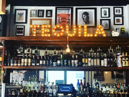 A bar shelf filled with various bottles, featuring a bright "TEQUILA" sign and framed artwork on the wall above.