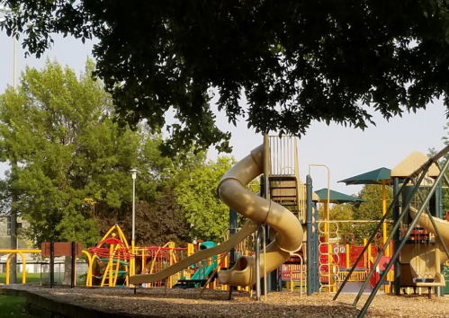 A playground featuring a large yellow slide, climbing structures, and green canopies surrounded by trees.