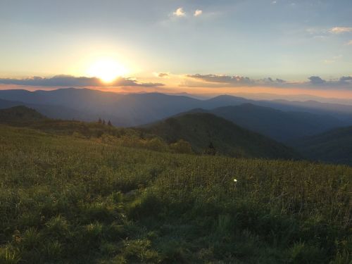 A sunset over rolling mountains, with a grassy foreground and soft clouds in a colorful sky.