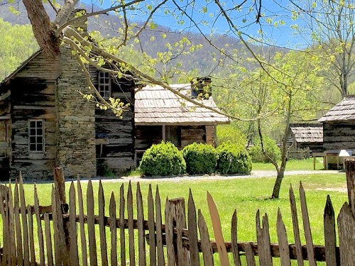 A rustic wooden house surrounded by greenery and a picket fence, set against a backdrop of mountains and blue sky.