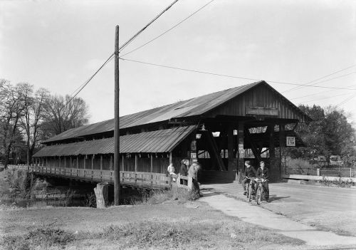 A historic covered bridge with people and bicycles nearby, surrounded by trees and power lines. Black and white photo.