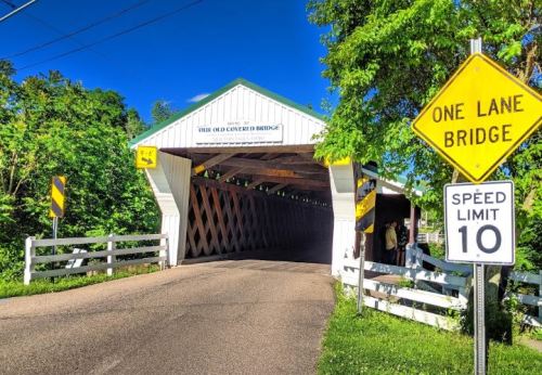 A white covered bridge with a sign indicating a one-lane bridge and a speed limit of 10, surrounded by greenery.