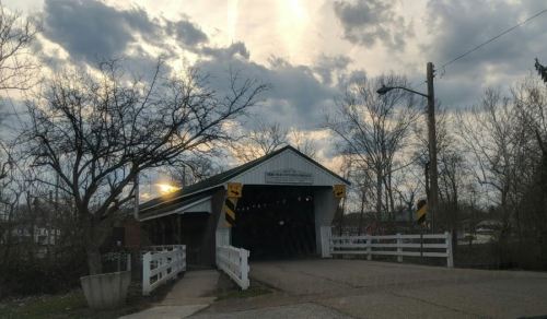 A covered bridge with a white fence, surrounded by trees, under a cloudy sky with the sun peeking through.