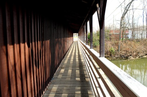 A wooden covered bridge with a long walkway, surrounded by trees and a river, casting shadows on the ground.