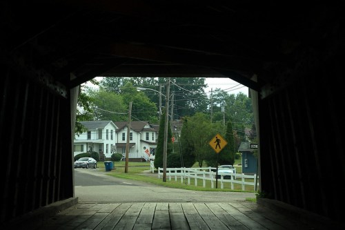 View from inside a covered bridge, looking out at a residential street with houses and a pedestrian crossing sign.