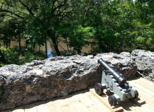 A historic cannon positioned on a wooden platform overlooking a river, surrounded by lush green trees.