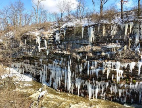 Icicles hang from a rocky cliff above a flowing river, surrounded by bare trees and a snowy landscape.