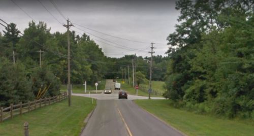 A rural road intersection with trees, power lines, and a cloudy sky in the background.