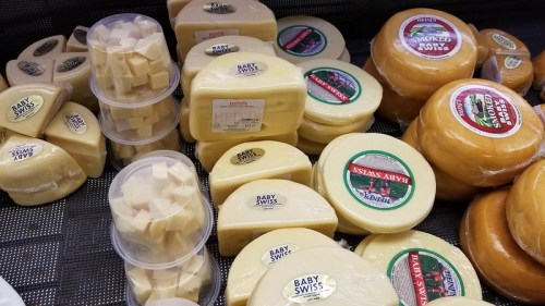 A variety of cheeses displayed in a market, including Baby Swiss and smoked cheese, in different shapes and sizes.