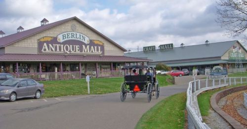 A horse-drawn carriage approaches the Berlin Antique Mall, with shops and cloudy skies in the background.