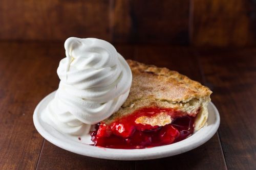 A slice of cherry pie with a flaky crust, topped with a swirl of whipped cream on a white plate.