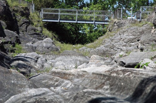 A rocky landscape with a bridge in the background, surrounded by greenery and trees under a clear blue sky.