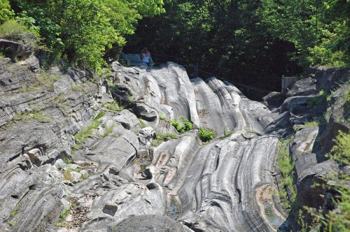 A rocky landscape with smooth, sloped surfaces and greenery, featuring a person in the background near a railing.