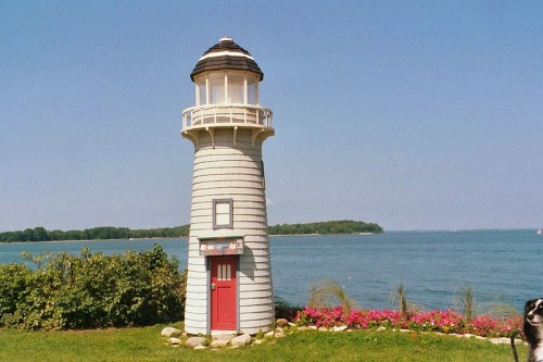 A small white lighthouse with a red door stands by the water, surrounded by greenery and colorful flowers under a clear blue sky.