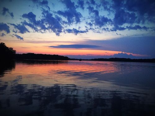 A serene lake at sunset, reflecting vibrant colors and clouds in the sky, surrounded by trees on the horizon.