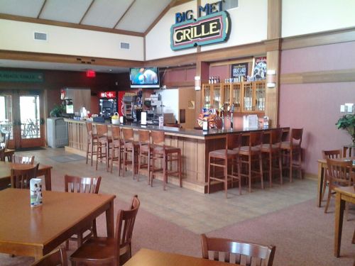 Interior of a restaurant bar area with wooden tables, chairs, and a counter displaying drinks and snacks.
