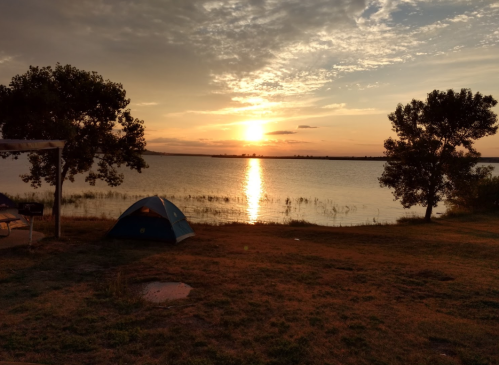 A serene lakeside scene at sunset, featuring a tent and trees silhouetted against the colorful sky.