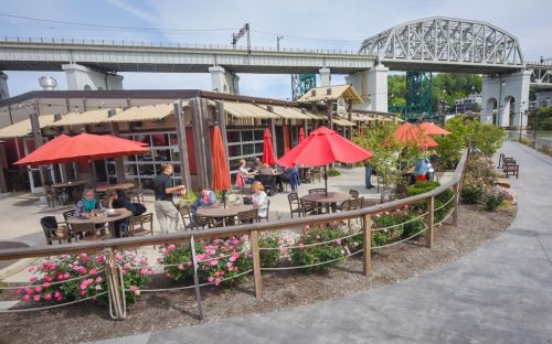 Outdoor dining area with red umbrellas, people enjoying meals, and flowers along a riverside path under a bridge.