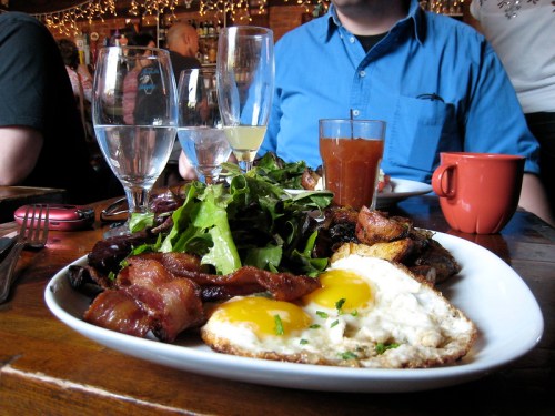 A plate of breakfast with eggs, bacon, salad, and potatoes, surrounded by drinks in a cozy restaurant setting.