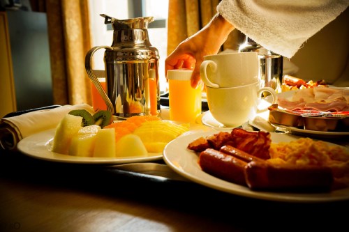 A breakfast tray with fruit, sausages, eggs, coffee, and juice, with a hand reaching for the juice.