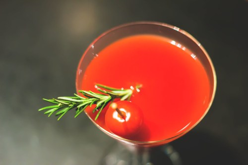 A vibrant red cocktail garnished with a cherry tomato and a sprig of rosemary, served in a martini glass.