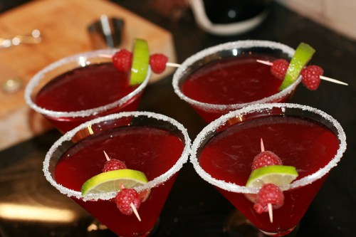 Four vibrant red cocktails with raspberries and lime slices, garnished with sugar rims, on a dark countertop.